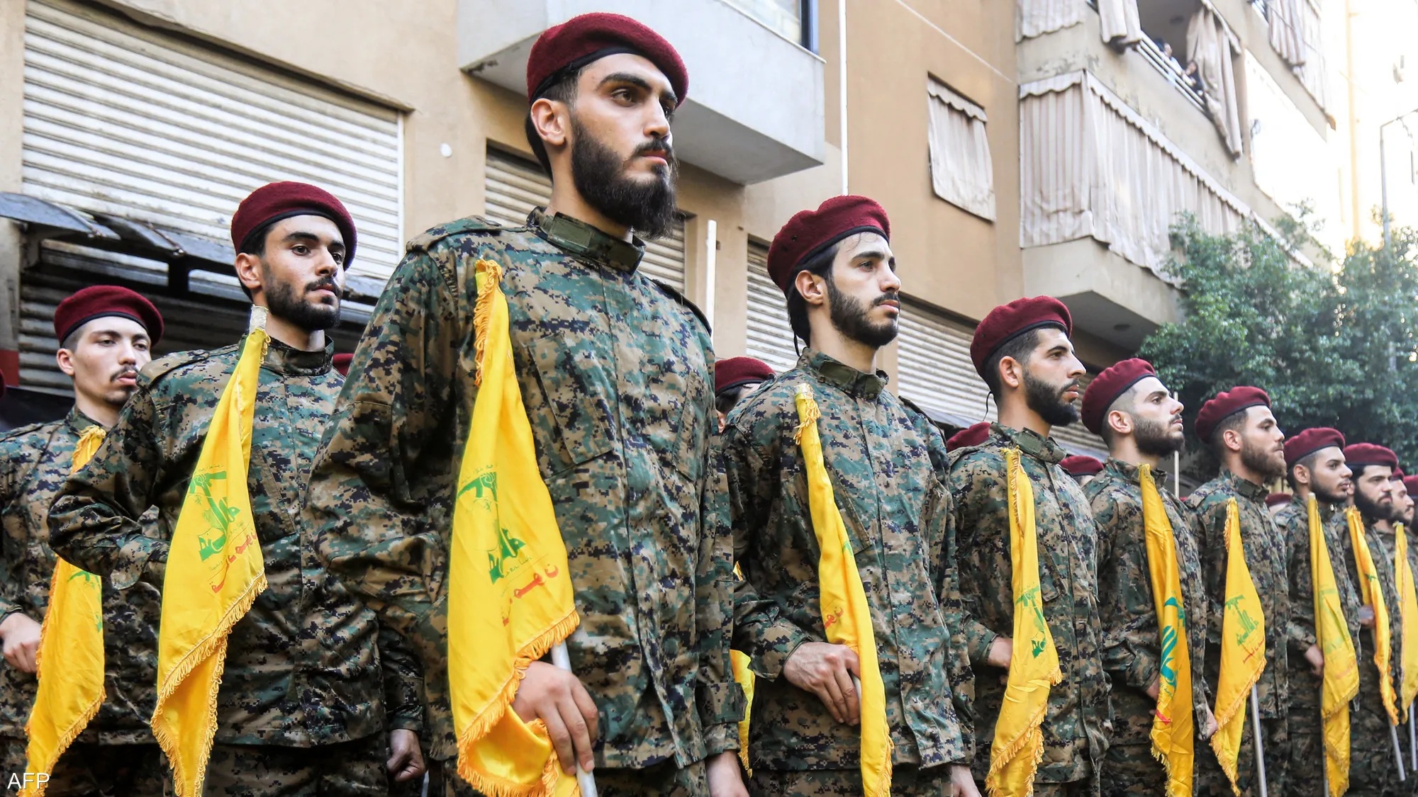Hezbollah fighters stand in formation during a public display, their uniforms and flags visible as they maintain a disciplined posture in an outdoor setting.