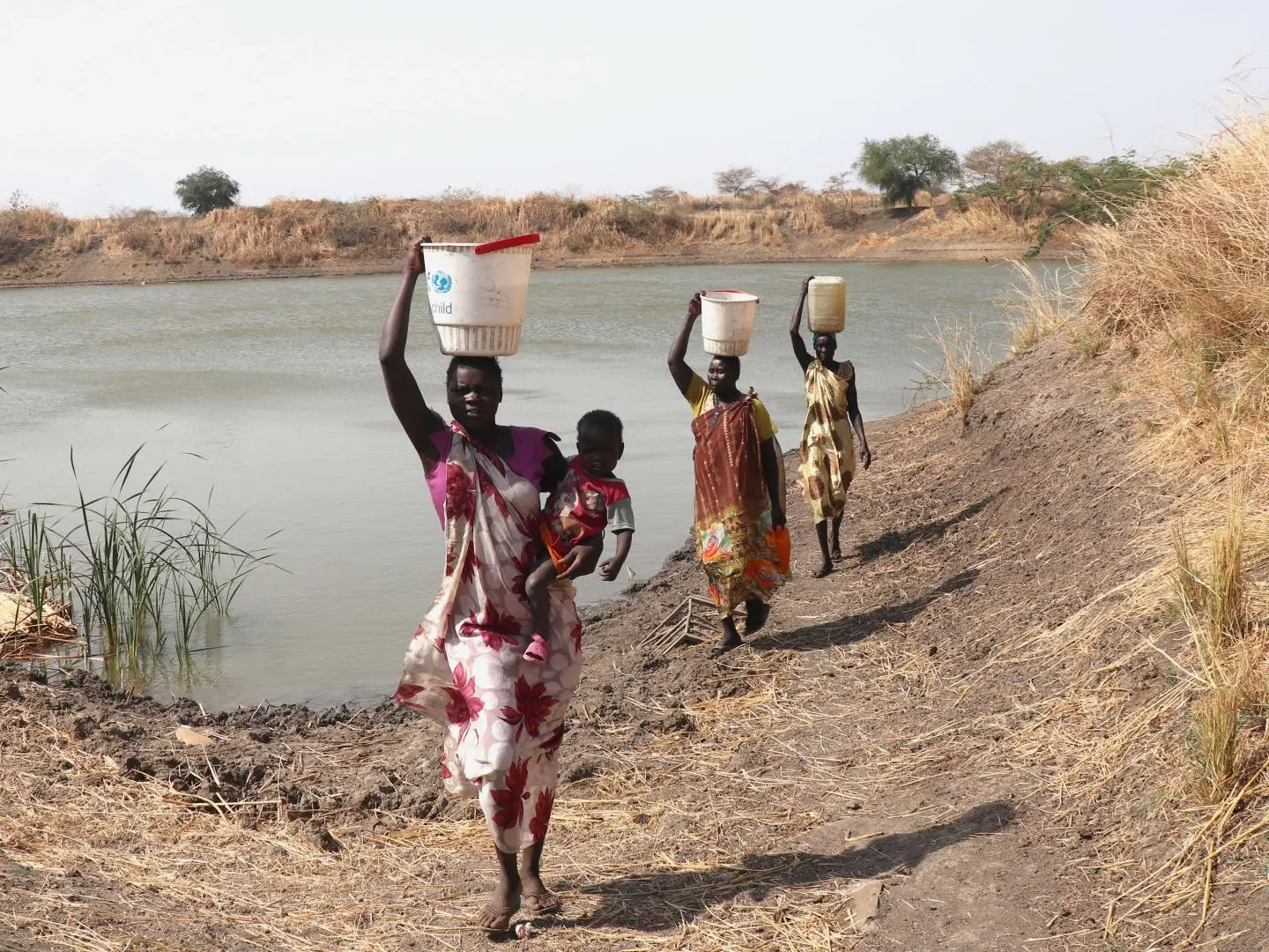 A woman carries a 20-liter water container on her shoulder after collecting water from a community source in South Sudan, where recurring conflict, displacement, and food insecurity continue to strain access to basic services.