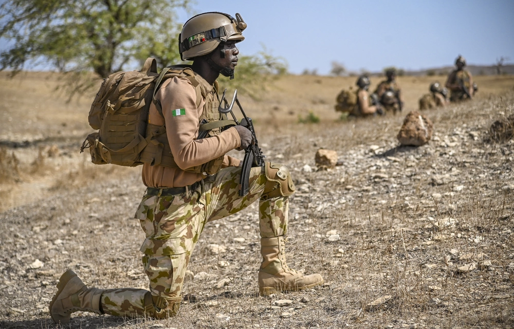 A Nigerian soldier crouches on one knee, gripping his AK-47 tightly as he scans the surroundings in a tense, alert stance during an ongoing security operation.