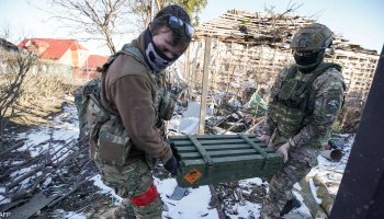 Two Russian soldiers carry and load ammunition at an active frontline during ongoing combat operations.