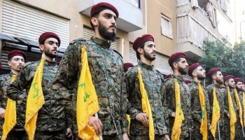Hezbollah fighters stand in formation during a public display, their uniforms and flags visible as they maintain a disciplined posture in an outdoor setting.