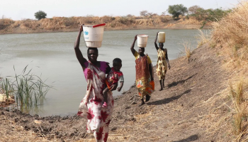 A woman carries a 20-liter water container on her shoulder after collecting water from a community source in South Sudan, where recurring conflict, displacement, and food insecurity continue to strain access to basic services.