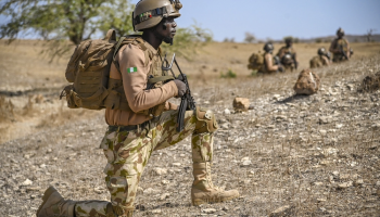 A Nigerian soldier crouches on one knee, gripping his AK-47 tightly as he scans the surroundings in a tense, alert stance during an ongoing security operation.